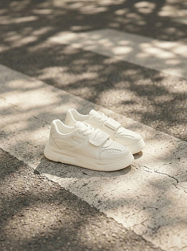 主体是用户提供的米白色魔术贴休闲板鞋，高度还原鞋子的所有细节，包括魔术贴设计、鞋面纹路、厚底结构，保持鞋身形态完全不变。参考参考图的户外斑马线场景和阳光树影的柔和氛围，将鞋子放在户外斑驳阳光照射的沥青路斑马线位置，光影为柔和的自然漫射光，树影斑驳洒落在地面和鞋身上，采用微侧15度视角拍摄，左右翻转参考图的光源方向，整体色调为温暖的奶油米色系，画面明亮通透，有自然的光斑流动感，符合国内电商种草氛围感。