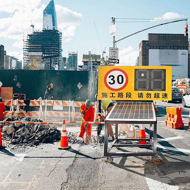 将抠好的施工路段测速警示标识放置到城市道路施工场景图的前方路边合适位置，自然融入画面，保持真实感