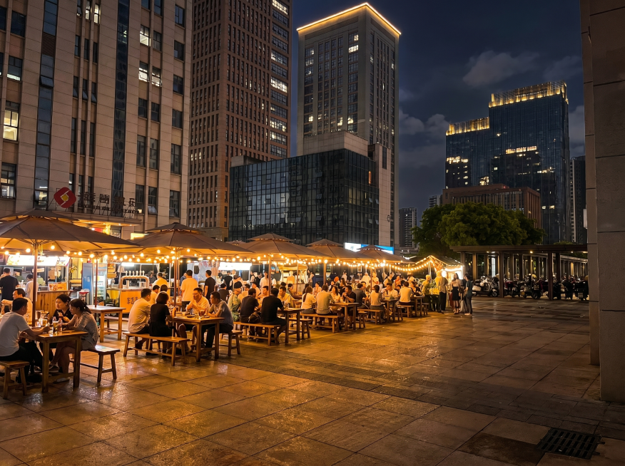 将第一张图片中的热闹啤酒广场场景合成到这张图片的空广场区域，去掉前景的四个石墩，整体改成夜晚灯光效果，保持整体透视和光线自然协调，保留建筑背景，替换前景广场部分为啤酒广场预览效果