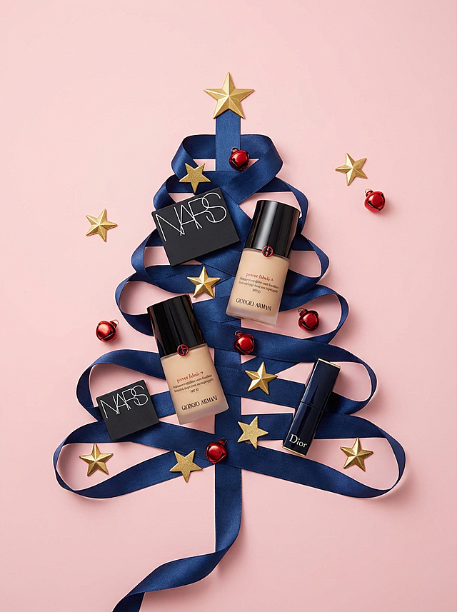 粉色背景上，深蓝丝带巧妙拼贴成圣诞树造型，点缀金色星星与红球，融合NARS、Armani、Dior等高端彩妆，呈现奢华喜庆的双色几何风节日视觉。