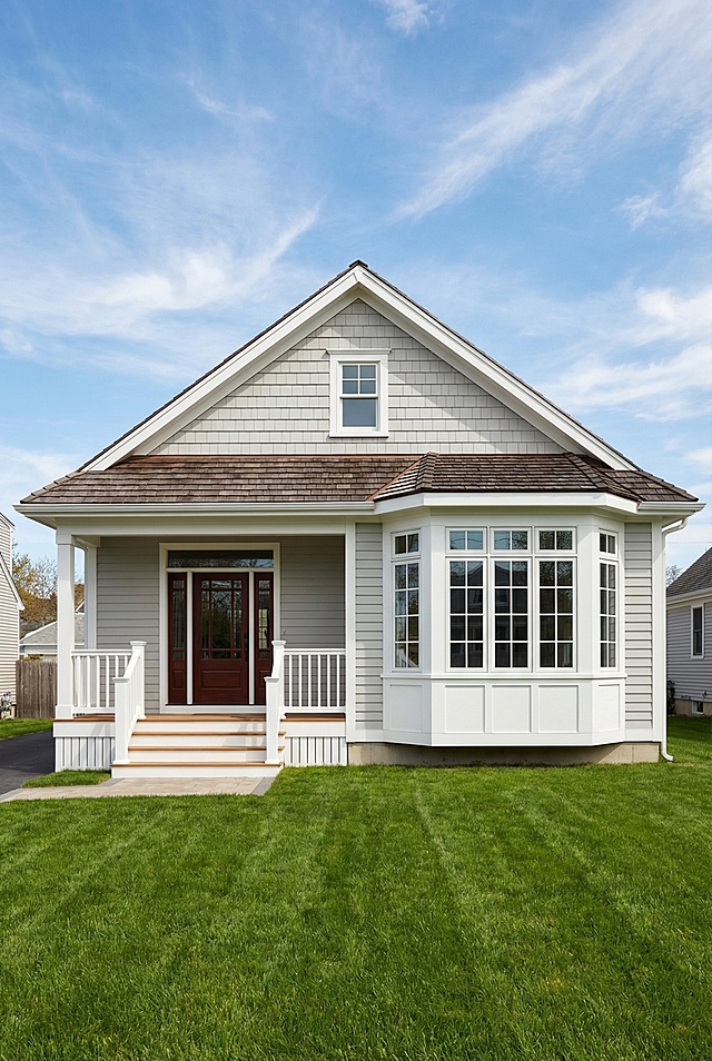 A single-story detached house with a light gray and white exterior sits under a clear blue sky with some wispy white clouds. The house features light gray horizontal siding on the lower half and shingle-style siding, also in light gray, on the upper gabled section. The trim around the windows, doors, and roofline is white or a very light beige. The main roof has a low pitch and is covered with what appears to be dark brown or gray shingles.

The front of the house has a small covered porch area on the left. Steps leading up to the porch have white railings and risers, with natural wood treads. The front door, slightly recessed under the porch roof, is a deep reddish-brown color with some window panels.

To the right of the porch is a prominent bay window structure with multiple panes, featuring a large central horizontal window flanked by two narrower vertical windows, all trimmed in white. Below the bay window, the siding is white.

The yard features a meticulously maintained, bright
