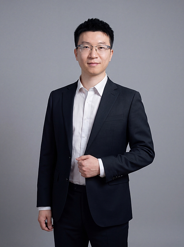 参考图中保留人物脸型、发型、年龄等特征生成职场形象照,标准焦段50mm,f/4光圈(f/2.8-f/4区间),平视视角(镜头与人物视线齐平);人物占画面70%,人物居中(居中构图),位于画面中心,为视觉聚焦点,入境范围头部及以下至臀部下方10cm(大半身像);左右留少量背景空间;头部与画面顶部保留5%空间;头部发型完整,头肩比例协调,人物结构准确;发质微微柔顺，微微蓬松。身穿深黑色精纺羊毛混纺西装外套,(平驳领设计,面料呈细腻微微哑光质感);内搭白色高支精梳棉衬衫;下装深黑色精纺羊毛混纺西裤;着装正式干练;专业稳重;身形匀称,姿态端正,身体挺拔。呈右手轻握西装前襟的自然姿态，肢体放松且自信,佩戴眼镜,去除多余配饰;简约微微中度灰色纯色背景(呈现均匀柔和质感),背景光线均匀柔和,强化色彩对比的高级感。极简干净;画质超高分辨率、细节清晰,现代精致感风格;肤质精细化处理;眼神直视镜头,目光坚定且澄澈,表情自然。嘴角上扬;肤质自然细腻,肤色健康均匀,皮肤状态清爽干净,保留真实皮肤细节｡正面柔光补光,4800K中性光,面部光比1:1.5｡中近景写实摄影,对焦清晰,面部雾面柔光,轻透裸肌感,高清保留皮肤细节｡分辨率4K,去除人物之外的所有元素。严格按照图片内容生成，画面比例3:4，高清出图，实拍大片。