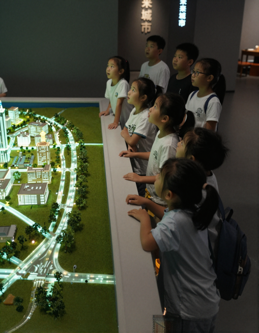 超写实室内摄影，浦东城规馆内，一群5-10岁孩子站在大型城市沙盘前，欣赏裸眼3D灯光秀。沙盘上光影流动，孩子们抬头观看，表情惊喜。室内光线较暗但面部有柔和补光，背景有"未来城市"字样。相机抓拍，电影感预览效果