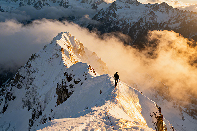 一名攀岩者在高耸的雪山峭壁上攀爬，周围是皑皑白雪，云雾缭绕在山峰间，阳光洒在雪面上泛着金光，画面充满冒险与震撼感