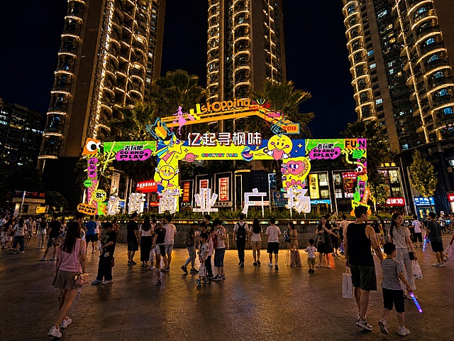 将这张商业广场照片改为夜景效果，前广场上增加中国购物休闲的游客，包含成人和儿童，凸显夜间商业氛围，广场上的彩色拱门装饰、文字、建筑、棕榈树等其他所有内容保持不变，画面整体保持真实摄影风格，夜晚灯光璀璨，商铺亮灯，氛围感强