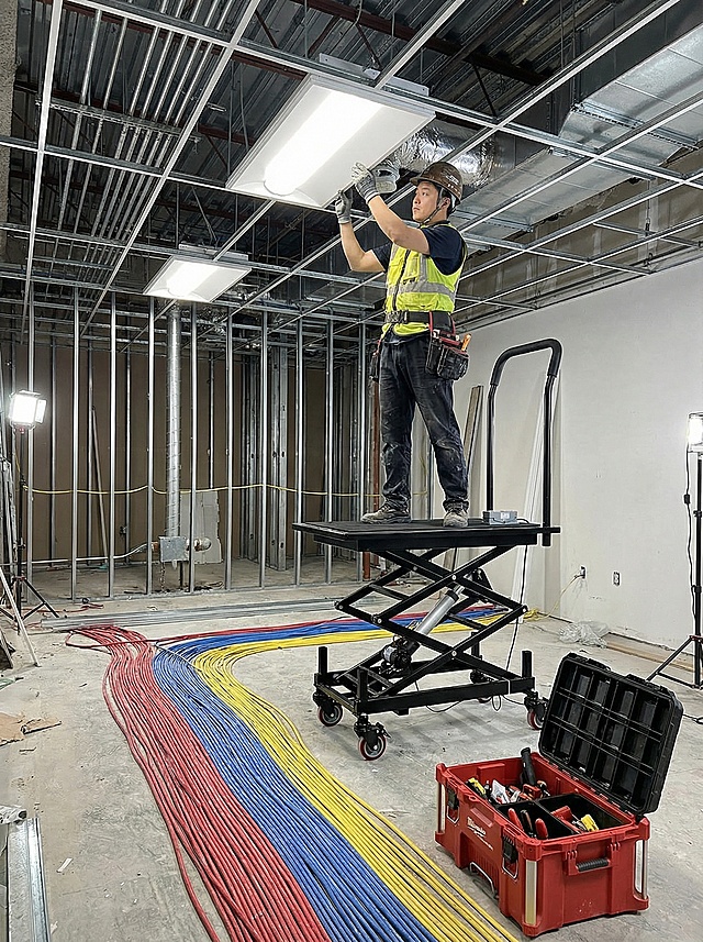 专业电工在商业建筑天花板上安装灯具，使用电动马凳精确升至一定高度，稳定承载其与工具，周围是铺设整齐的线缆和工具箱。   比例3:4 不要加文字 人物为中国人