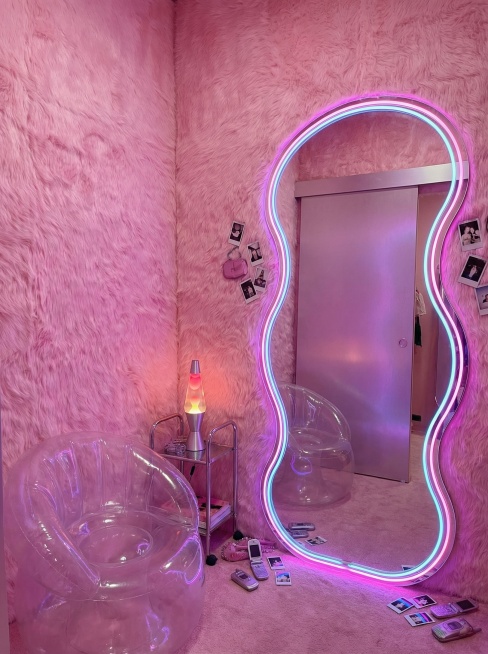 豪华又好玩的Y2K风格内衣店试衣间。墙壁覆盖着柔软的粉色人造皮草。镜子不是标准的矩形，而是一个巨大的、变形虫形状的全身镜，带有发光的霓虹边框。角落里放着一把小巧的透明充气椅。灯光柔和且显瘦，由一个放在预览效果