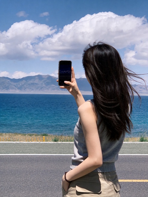 女生游客在青海湖边的背影自拍，女生站在湖边公路旁，背影朝向蓝色湖泊和远方山脉，头发被风吹起，穿着简约休闲装，画面开阔治愈，蓝色湖泊搭配白色云朵，自然真实游客打卡照风格，竖版适合小红书配图，画质清晰预览效果