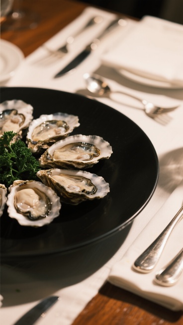 写实风格美食摄影，特写镜头，新鲜撬开的生蚝，肉质饱满富有光泽，壳上带有自然的灰褐色纹理，黑色圆形餐盘中堆叠的生蚝，旁边点缀着翠绿的欧芹，背景变换为高档餐厅的木质餐桌，搭配白色餐布和精致的银色餐具，柔和的暖色调侧光，突出生蚝的鲜嫩质感和水润光泽，整体营造出优雅奢华的用餐氛围，画面清晰锐利，色彩真实自然，细节丰富。预览效果