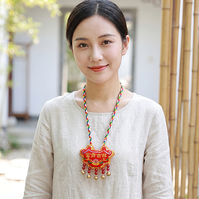 摄影写真，一位美女胸前挂着参考图中的端午香包，香包为红色锁形刺绣，上有黄色文字“富贵吉祥”和粉色花朵刺绣，下方垂挂红色珠子和金色铃铛，搭配彩色编绳挂绳，画面整体清爽氛围感，高质量质感，1:1构图