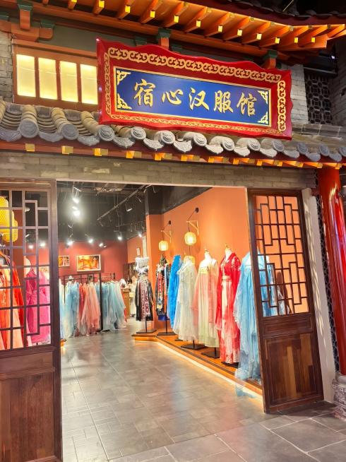 修改图片中的招牌文字，将原招牌的“国潮汉衣馆”替换为“宿心汉服馆”，保持招牌原有红底金纹蓝底金字的中式国风设计风格，字体为传统书法字体，整体协调美观，保持原图片其他内容不变预览效果