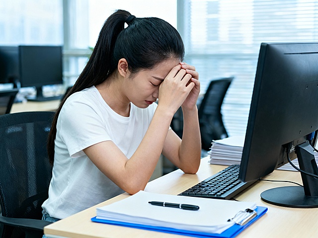 一位年轻亚裔女性坐在办公室电脑前，双手交叠按在额头上，头低垂，双眼紧闭，表情疲惫痛苦，黑色长发扎成高马尾，穿着纯白色短袖T恤。她坐在黑色办公椅上，面前浅色木桌上有黑色电脑显示器、键盘、堆叠的文件和蓝色文件夹，文件夹上放着一支黑色签字笔。背景是模糊的办公室环境，有其他电脑和办公桌椅，自然光从百叶窗透入，整体色调明亮偏冷，氛围压抑，8K超高清，写实摄影风格，细节清晰，专注于人物的疲惫姿态和手部动作。