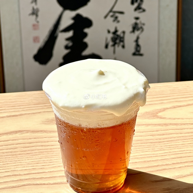 特写镜头下的一杯刚做好的奶茶，顶部覆盖着厚实绵密的芝士奶盖，奶盖边缘微微下塌，杯壁凝结着晶莹的水珠，折射出柔和的光线。奶茶杯放置在浅色橡木桌面上，木纹清晰可见。背景虚化处理，隐约可见一幅中国书法画，增预览效果