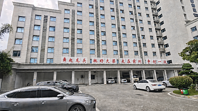 将建筑上方的四组红色口号标语（舞起龙头、挑好大梁、走在前列、争创一流）整体向右平移一个车库门的距离，保持四组口号之间的缩小后的间距不变，保持白底红字的样式和字体不变，建筑、车辆、树木等其他所有画面元素保持原样不变，自然真实，符合透视