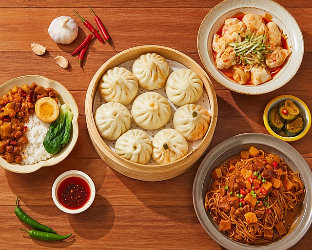 超写实美食摄影，90度垂直俯视拍摄。

主体：包子严格参照参考图产品样式，完整保留褶皱、竹蒸笼、轮廓、结构、比例、材质、颜色产品特征，包子放置图片正中央；其他餐品卤肉饭、干捞面、肠粉、腌黄瓜、红辣椒、大蒜错落有致平铺在图片两侧，保留原有餐具和产品细节，所有产品色泽自然新鲜，无变形扭曲。

构图：产品均匀分布，包子为视觉中心，俯拍平铺构图。

风格：国内电商美食风格，真实有食欲。

场景：原木餐桌面，干净简洁。

陈列：
①图片正中央，蒸笼盛放的包子，竹制蒸笼米黄色；
②图片左侧，盛放卤肉饭的黄边陶瓷碗，搭配青梗菜和卤蛋；
③图片右下方，盛放焖肉干捞面的灰色陶瓷盘；
④图片右上方，盛放肠粉的米黄色陶瓷碗，搭配黄瓜丝；
⑤图片最右侧，小碟装腌制黄瓜。

配色 / 材质：米黄色、原木棕、红色、绿色、白色；原木、陶瓷、竹制、面食、新鲜蔬菜。

光影：明亮暖调主光从上方均匀照射，产品呈现自然清晰阴影，面皮纹理和酱汁光泽自然，激发食欲。

严格锁定宽高比为 1.25:1，4K超高清，产品细节清晰。