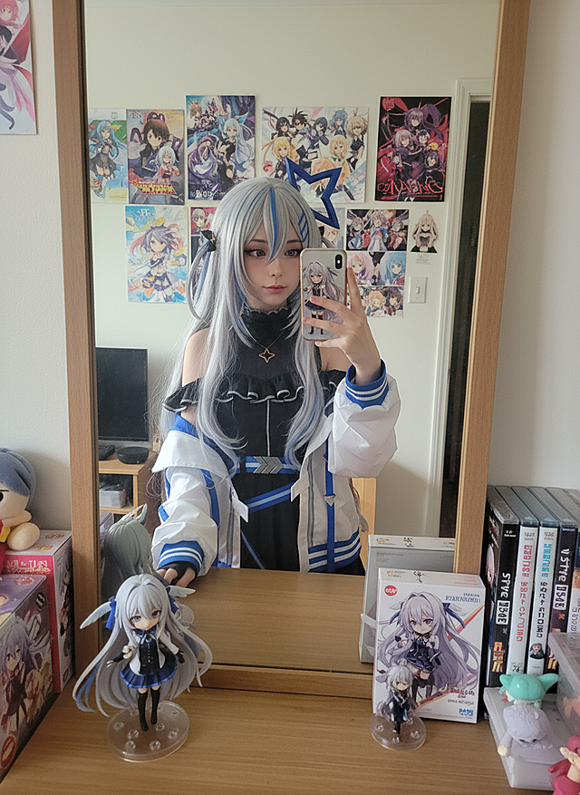 cosplayer真人照片，根据该图生成一张现实生活中扮演该角色的写实风格，人物所在的写实场景与原作相对应，人物一只手抬起拿着手机对着镜子正在自拍，另一只手靠近镜头托着带有与该cosplayer照片相对应的玩偶手办。