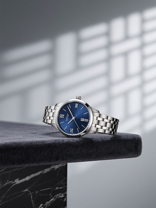 一块银色圆形表盘的男士机械腕表，表盘为深邃的蓝色，搭配银色指针和罗马数字时标，银色金属表带设计精致，腕表优雅地斜靠在铺有深灰色丝绒的黑色大理石台面上，背景是柔和的浅灰色墙面，墙上投射着细密的几何图案光影，整体画面呈现出现代简约的高端质感，光线柔和，阴影细腻，突出腕表的金属光泽和材质细节，营造出静谧而奢华的氛围。