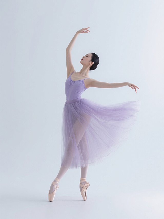 一位身着淡紫色纱质芭蕾舞裙的东方女性舞者，在纯白色背景中展现优雅的阿拉贝斯克舞姿，左腿向后伸直，右腿支撑身体，双臂舒展如天鹅展翅，裙摆随动作自然飘逸形成优美弧线，舞者面容沉静专注，肌肤细腻有光泽，整体画面采用高清晰度摄影风格，光线柔和均匀，突出舞者轻盈灵动的姿态与纱裙的通透质感，无任何文字及装饰元素。