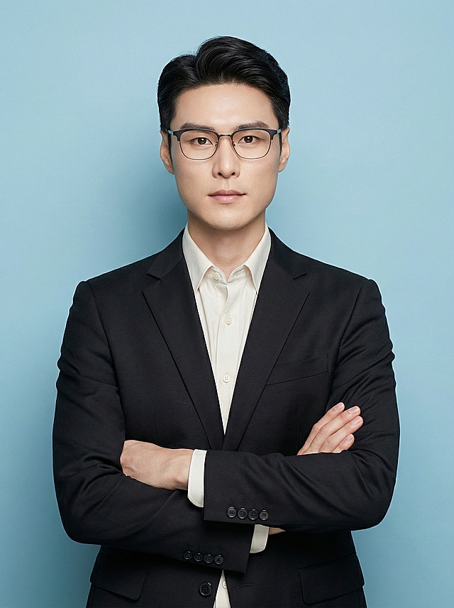 模仿参考照片风格，生成一位男教师证件照，人物正面站立，双臂交叉于胸前，穿着黑色西装外套内搭米白色衬衫，浅蓝色纯色背景，柔和均匀的商业人像布光，突出人物面部
