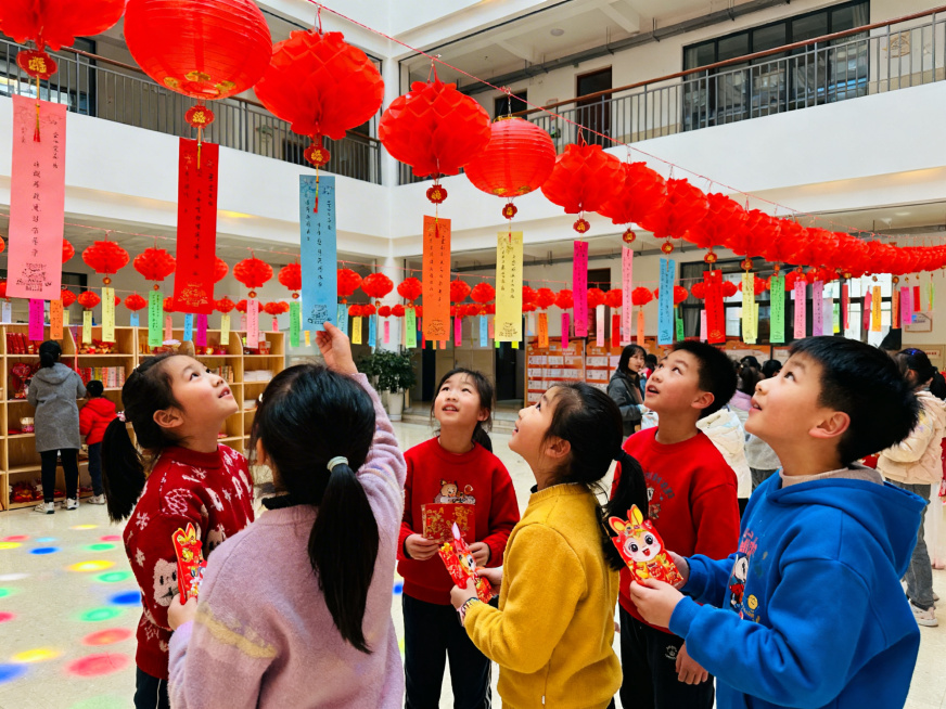 真人实拍风格，少年宫楼下的元宵灯会场景，一群孩子围在灯谜展架前，有的仰头读灯谜，有的兴奋地拿着猜对灯谜兑换的小礼物，现场氛围热闹欢快，挂满红色灯笼和彩色灯谜条预览效果