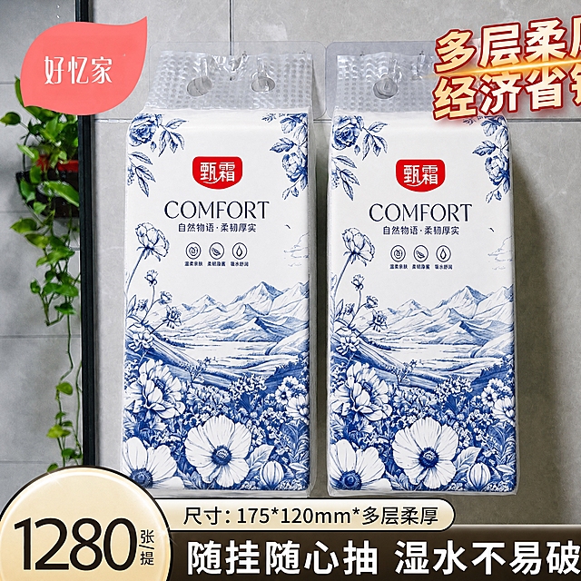 将第一张白底图（甄霜蓝白青花风格悬挂式面巾纸包装，包含红色'甄霜'品牌标识、'COMFORT'英文、'自然物语·柔韧厚实'中文、三个功能图标、蓝白山水花卉图案）的完整包装设计图案，精准迁移替换到第二张图中两个悬挂式面巾纸的包装表面。保持第二张图的场景环境（灰色瓷砖墙面、左侧绿植、顶部挂扣、底部文字和价格标签等所有背景元素）完全不变，包装的透视角度、光影、褶皱、立体感与原图一致，图案贴合包装曲面自然真实，蓝白青花风格完整呈现，品牌文字和图案元素全部保留。