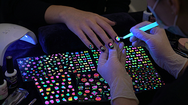 保持原图中各类夜光美甲贴纸的图案和发光特征不变，展示欧美女人做美甲时使用这些夜光贴纸的过程，贴纸在暗光环境下绚丽发光，超清美妆摄影，16:9构图