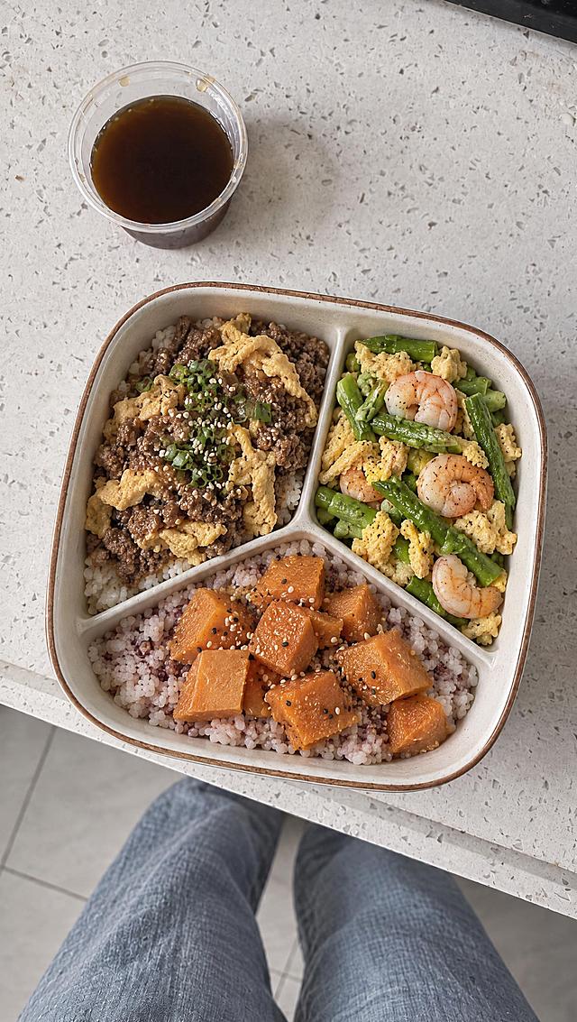 俯视视角，白色带棕边的方形便当盒里装着牛肉末煎蛋饭、红薯饭、芦笋虾仁炒蛋三格。旁边有一小杯深色酱汁。背景是浅灰色带细斑点台面，画面下方能看到穿着蓝色牛仔裤的腿。低饱和度ins风滤镜，柔和自然光，暖灰色调，美食摄影，真实质感