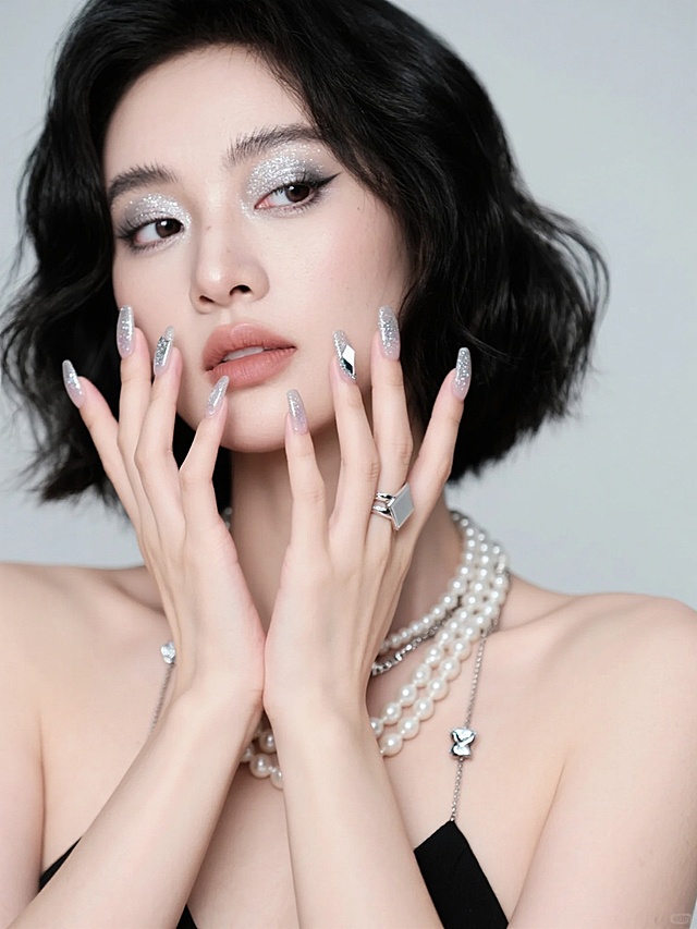 一位女性双手捧脸的时尚人像摄影，黑色波浪短发，银白色闪亮眼妆，自然唇色，佩戴多层珍珠项链与银色戒指，背景为浅灰色。双手指甲为精致的美甲设计，采用透明底色搭配银色闪粉渐变与不规则几何银色亮片装饰，指甲形状为优雅的方形甲，整体风格现代、时尚、具有高级感，光线柔和，突出人物面部与手部细节，营造简约而奢华的氛围。