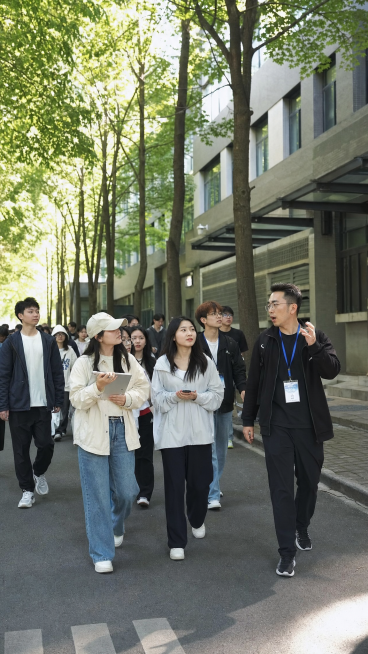 一群学生在大学校园里进行实地参观考察，大家跟随领队行走在校园的林荫道路上，周边是高校教学楼建筑，整体氛围青春活力，真实摄影风格预览效果