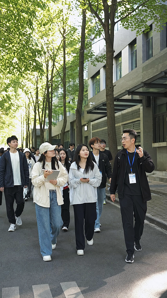 一群学生在大学校园里进行实地参观考察，大家跟随领队行走在校园的林荫道路上，周边是高校教学楼建筑，整体氛围青春活力，真实摄影风格