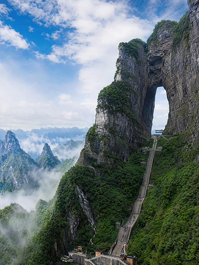 天门山天门洞全景，巨大的天然穿山溶洞高悬于千米峭壁之上，气势磅礴雄伟壮观，999级石阶天梯直通天门洞，周围云雾缭绕，远处是连绵的张家界喀斯特峰林，蓝天白云，震撼人心的自然奇观，高清摄影风格，竖版构图