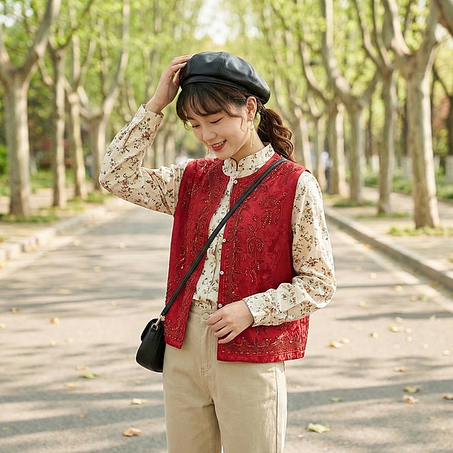 同一个长相甜美的年轻东亚女生，年龄20-25岁，身穿图中这件红色提花烫钻马甲，搭配浅卡其色直筒休闲裤和米白碎花立领衬衫，上衣不塞下装。女生留高马尾，发尾烫出自然弧度，额前留碎刘海，头戴黑色皮质贝雷帽，佩戴金色长款耳环，斜挎黑色小方包。清透自然淡妆，嘴角噙着温柔笑意。女生做单手扶帽摆拍动作，这是一张全身人物特写图片，从右侧45度角度拍摄，背景为春日公园林荫道，两侧是嫩绿梧桐树，地面散落少量落叶。自然柔和的日间散射日光，整体暖调通透，没有生硬阴影，色彩温润自然，人物对焦精准，皮肤质感真实细腻，整体充满松弛慵懒的春日日常感，生活气息浓郁