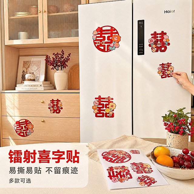 [主图-1] 镭射不干胶喜字贴——真实生活场景应用首图，唯一任务：让买家一眼看清这是什么贴纸、贴在哪里、贴完什么效果。

[统一基调] 暖调家居实拍风格；背景主色为暖白与浅木色，标题字色为喜庆正红，辅助文字为深灰，强调色取镭射红金反光；只用一种无衬线黑体，主标题字重最大最粗，辅助文案字号缩小一半以上；不使用厚重色块底板与大面积通栏边框，仅在下角加轻微半透明浅底提升可读性。

[画面] 现代简约家庭的厨房餐区实景：正前方一台白色双开门冰箱，左侧连接浅木色餐边柜与玻璃吊柜，冰箱门与柜门上分别贴着多枚大小不一的红色镭射不干胶喜字贴，包含圆囍与方囍两款，图案带粉橙卡通花朵与"love"小气泡，贴纸表面有镭射红金细闪，边缘服帖无气泡，分别贴在冰箱门中上部、柜门中部与下方抽屉，错落但不杂乱；台面右侧放一叠尚未撕下的喜字贴离型纸整版和一枚刚揭下的小喜字贴，旁边有浅色亚麻布、一小盆红果绿植与装着水果的陶瓷盘；画面右下角伸入一只女性的手（仅露出手部，涂淡粉指甲），食指与拇指正轻轻抚平冰箱门上刚贴好的一枚小喜字贴，光线自然、指节结构正常、无多余手指。产品外观、闪粉颗粒、卡通花朵与"love"气泡图案、颜色与输入图完全一致，不改变款式结构。

[光影质感] 上午自然光从左侧窗户斜射进来，柔和不均匀，冰箱门与柜门上有真实高光与柔和接触阴影，喜字贴随门板弧度产生轻微反光；整体为手机实拍质感，保留轻微噪点、真实材质颗粒与生活痕迹，不做过度磨皮、过曝与夸张锐化。

[配色] 暖白与浅木色作环境主色，红色喜字贴为视觉焦点，红金镭射反光为强调，绿色小盆栽与红果点缀。

[文字版式] 左下方空白处排版：第一行大号正红标题"镭射喜字贴"，第二行深灰小字"易撕易贴 不留痕迹"，第三行更小字"多款可选"；全部横排，不遮挡产品、贴纸与手部。

[输出要求] 1:1 正方形，文字清晰可读、产品清晰锐利；负向：不要平台Logo与水印、不要错字乱码、不要贴纸变形或图案错乱、不要悬浮与硬切边缘、不要白底残留、不要多余手指与畸形手、不要假花假材质、不要AI感塑料感与过度磨皮。