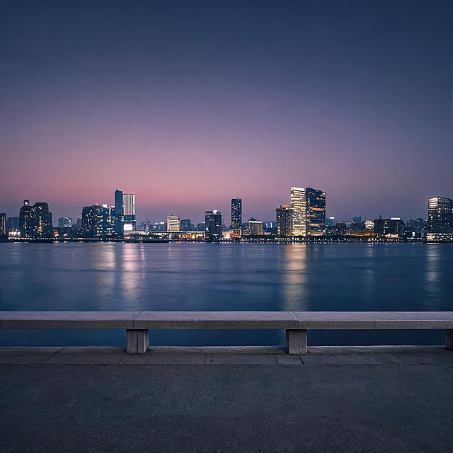 写实城市夜景摄影，傍晚暮色时分。临江的水泥护栏台面空无一人，安静地横在画面前景。面前是宽阔平静的江面，对岸是灯火璀璨的城市天际线，高楼大厦错落林立，各色暖黄与冷白灯光星星点点，柔和倒映在深蓝色水面上。天空从上方的深紫灰色向下渐变至靠近地平线的粉紫色暮色。整体色调偏冷蓝紫调，氛围空旷宁静、文艺治愈，电影感光影，景深效果，画面干净，无任何文字、无人物。