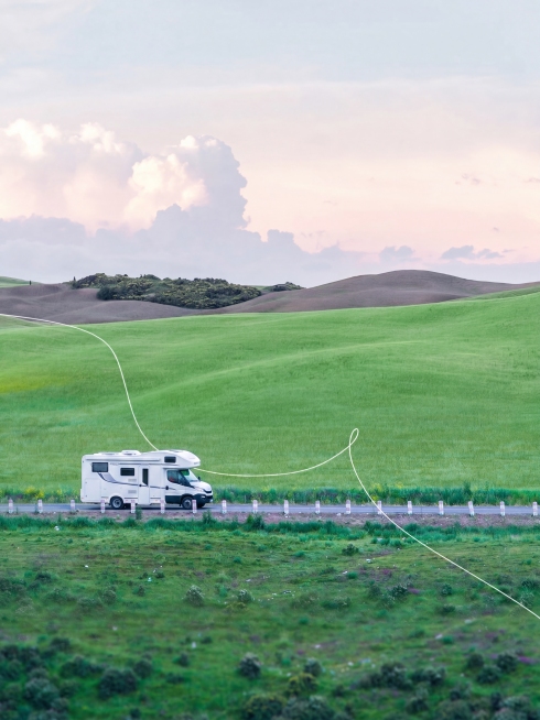 将图片中的红色大巴车替换为一辆IVECO C型房车，保持房车在同样的位置，同样的行驶方向，保持和原图相同的比例，保留周围的绿色草原、道路、天空和白云等所有原有景物不变预览效果