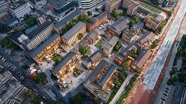 在这张文创园区鸟瞰效果图的建筑外立面和入口处，增加清晰可见的酒店、咖啡馆、书店、手作工坊等店招和招牌，如“酒店”“咖啡”“书店”等字样与简洁标志，店招设计符合旧改文创园区风格，材质自然（木质、金属或亚克力发光字），灯光温暖；保持原图的拍摄视角、机位、取景范围和构图完全不变，其他建筑、广场、绿化、人物活动场景保持不变