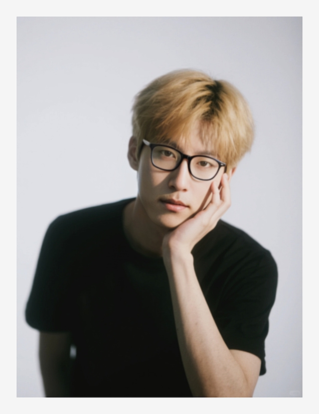 一个男生，金色头发，黑色短袖，单手托腮，脸部强曝光光线，正脸特写，低饱和度，加强阴影，白色背景。