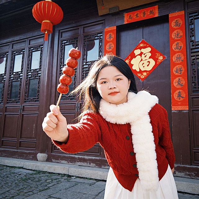 户外中式庭院新春写真头像(画幅比例1:1)画面聚焦人物;气质甜美治愈+复古胶片风格人物主体:冷白皮细腻通透肤质;橘红色哑光唇釉+浅棕自然眼妆,嘴角微微上扬,眼神明亮带笑意,注视镜头,气质灵动活泼､温暖亲和环境与构图:中景,三分构图(人物占画面85%);平拍偏微仰视角(拍摄距离1.5米);中式古建木门背景,贴红底金福字､竖版春联,上方挂红色灯笼,地面为青石路;着装与动作:穿红色羊毛混纺麻花纹理针织开衫,搭配净白色蓬松毛绒围巾;净白色裙装;单手举6颗山楂的糖葫芦串伸向镜头(极强的透视感,前景元素虚化朦胧效果),身体微微前倾,俏皮灵动,发丝被微风吹拂;光影与色彩:侧逆光午后自然光(4000k暖光),环境反光补光柔化面部阴影,发丝形成金色轮廓光;主色调大红色+米白色,暖调复古胶片色调,色彩高饱和柔和画质与氛围:超高清分辨率,去除人物之外所有元素和配饰;中等锐度,添加轻微胶片颗粒感(iso200);复古胶片滤镜(曝光+5,对比度,3,饱和度+10,颗粒+8),强化温暖治愈的新春氛围｡