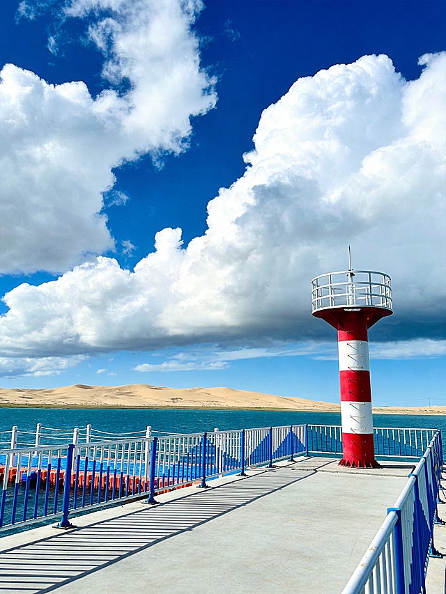青海湖二郎剑景区，蓝色湖面停靠红白相间的灯塔，大片蓬松棉花糖积雨云漂浮在深蓝色天空，远处沙丘，深蓝湖水清澈，光影通透，治愈清新的西北风光，高饱和色彩