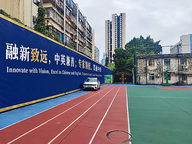 保持这张照片的整体场景不变，只在左侧深蓝色喷绘布围挡上添加致远新校训文字，文字沿围挡横向排版、覆盖围挡大部分长度，白色与金色大字，清晰端正、无错别字：中文第一行“融新致远，中英兼善；专家领航，贯通中西”，下方一行较小英文“Innovate with Vision, Excel in Chinese and English; Experts Lead, East Meets West”；不要改动操场、篮球场、白色汽车、大门和建筑。