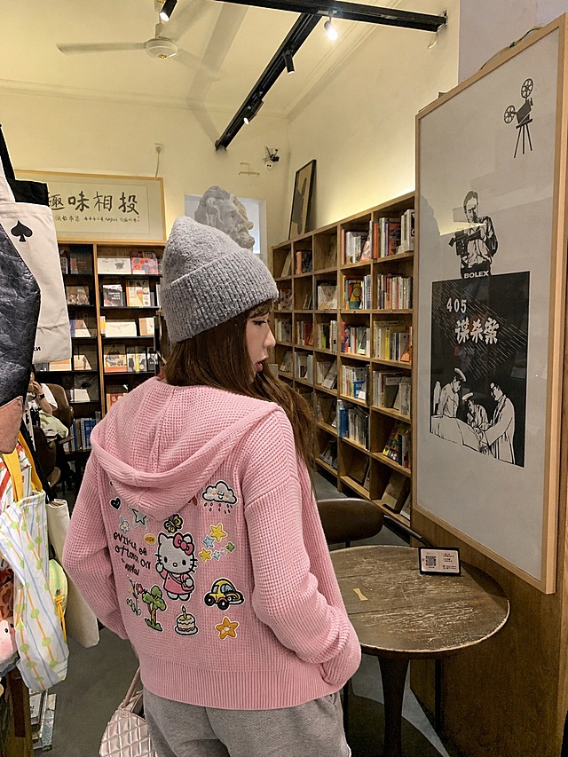 将图1和图2中的粉色华夫格连帽拉链卫衣穿到图3的模特身上，保留图3人物的五官、脸型、发型、姿势、身体姿态完全不变，人物长相不做任何改变。衣服为修身版型，正面是粉色华夫格针织连帽拉链开衫，背面有Hello Kitty卡通刺绣贴布图案及星星、云朵、小车、蛋糕等装饰元素，衣服的花色、纹理、面料、图案位置严格复刻参考服装。真实人像摄影，自然光，皮肤质感真实，高清写实，柔和环境光影，衣服贴合身体，服装褶皱自然，无水印，无文字，无logo，无畸形肢体，画面干净，保留原书店背景环境。
