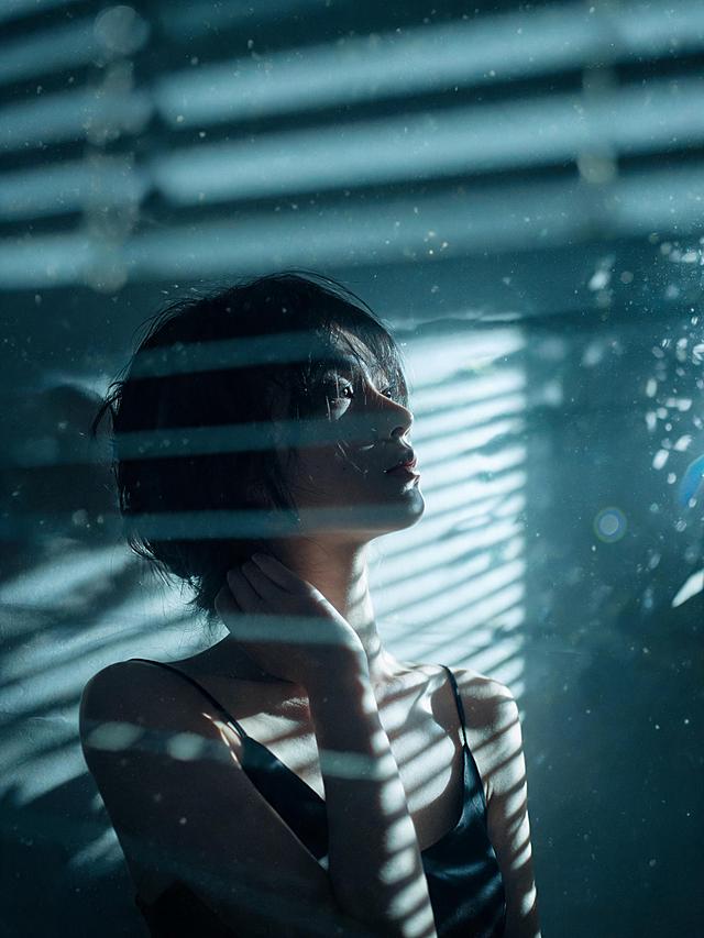 生成光影切割百叶窗与树影主题写真，王家卫式电影叙事风格，青灰色冷调滤镜，高噪点粗颗粒胶片质感，低饱和度，迷离暧昧破碎的故事感；强烈的局域硬光聚光灯投射，百叶窗几何条纹光影在人物脸部与身体形成锐利切割线，明暗交界分明，暗部深邃死黑，镜头微眩光，前景百叶窗叶片虚化遮挡；清透哑光肤质，真实皮肤纹理，细腻毛孔，冷调白皙；黑色短发微乱，几缕发丝落在额前；不拍全脸，半张脸隐没在阴影中，只露出鼻梁、唇线与下颌的锐利轮廓，侧脸微抬，眼神望向画面外，表情沉静疏离，单手轻搭在颈侧，手指修长；身穿简约黑色吊带背心，丝滑哑光面料，肩带纤细；暗调室内极简空间，深灰墙面，一盏硬光从侧方打入，空气中有细微尘埃在光束中浮动，树影与百叶窗条纹交织投影在墙面与人物身上；前景百叶窗叶片虚化形成框架构图，中景人物侧身居于画面三分之一处，偏心构图，截图感，85mm焦段，浅景深；分辨率4k，电影级光影质感，商业人像摄影级别，画面比例3:4