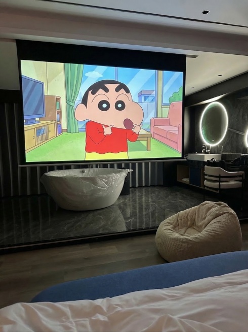 把投影幕布的投屏界面替换为蜡笔小新的动画画面，在木质地板靠近床的位置放一个米白色的懒人沙发，保持整体暗调轻奢风格协调统一预览效果