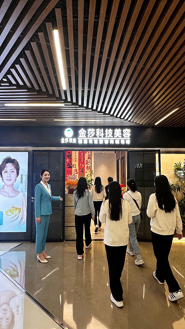 以图一实景门店场景作为主体完整背景，还原场景细节，一位女性专业美容顾问身着和图二款式版型、配色高度一致的统一浅蓝色拼白色翻领西装工作服，站姿优雅站在门店门口，不看镜头，面带温柔亲和笑容热情看向顾客引领到门店内，6位身着休闲服装的女顾客零零散散有些距离的走进店内，人物神态自然生动，画面质感超清细腻，64K超高清画质，电影级光影氛围感，写实真人风格，竖版构图，严格9:16比例，细节拉满，实景真实拍摄感