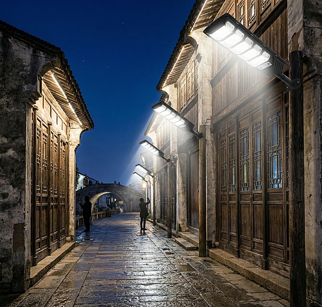 夜幕初降，华灯初上。白色的灯光从古老的墙壁上洒下，既照亮了脚下的青石板路，又温柔地洗亮了古建筑的屋檐和木窗。三到五盏灯在画面中形成有韵律的光线引导，远方是深蓝色的天空和古桥的剪影。一两位游客的剪影正驻足观赏。