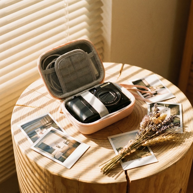 将背景替换为浅木色原木桌面，周围散落几张拍立得相片和一束干花，添加柔和的百叶窗斜射阳光，在包身上形成明暗相间的条纹光影，整体呈现温暖治愈的胶片感，保留相机包收纳着白色线缆和黑色小相机的状态预览效果