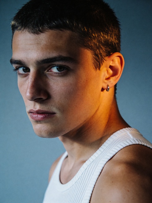 生成25岁欧美男生模特写真，冷调极简时尚风格，胶片蓝灰调质感，低饱和蓝灰背景搭配暖橙肤色，轻微颗粒复古电影质感；侧光硬阴影打光，冷暖对比，暖光打在面部冷色背景，高对比度突出面部明暗层次，发丝带有边缘光预览效果