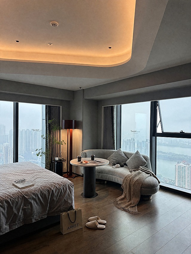 城市江景酒店弧形沙发房，保持整体色调不变，画面真实自然协调，增强生活感和入住感，素人感，精致好看，不要AI感，不要太假。增加更多素人感摆件物品，物品更丰富，有生活感和氛围感，小红书高端酒店氛围，真实感，4K高清，不拼图。小圆桌上放两杯咖啡、一盘马卡龙、一本杂志，沙发上搭一条毛毯和抱枕，床上放一本书和一副眼镜，落地灯旁边放一个小边几放着香薰，地上放一双拖鞋和一个购物袋，增加小绿植、护肤品、耳机等生活细节。