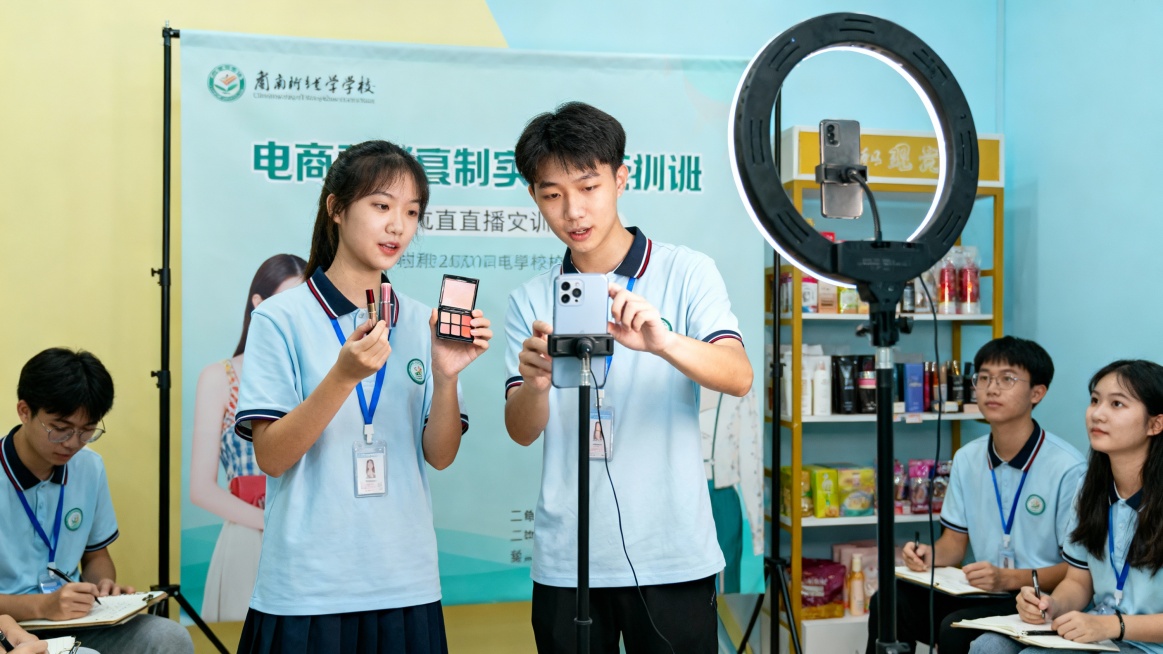 电商直播实训区，学生两两协作，一人手持美妆 / 服饰样品讲解，一人操作手机调整直播画面、回复评论，背景是学校定制的直播背景板，旁边立着补光灯和商品陈列架，其他同学在旁观摩记录，清新明亮风格，充满学习氛预览效果