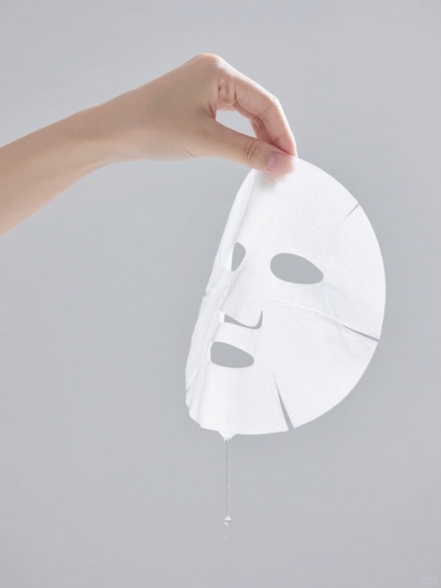一只女性的手轻柔地捏着一张白色的面膜，面膜质地轻薄、半透明，边缘自然垂落，面膜上的精华液正缓缓滴落，背景为纯净的浅灰色，整体画面呈现出简约、干净、专业的护肤品广告摄影风格，光线柔和均匀，突出面膜的细腻质感和水润状态，营造出宁静、舒适、专注于肌肤护理的氛围。预览效果